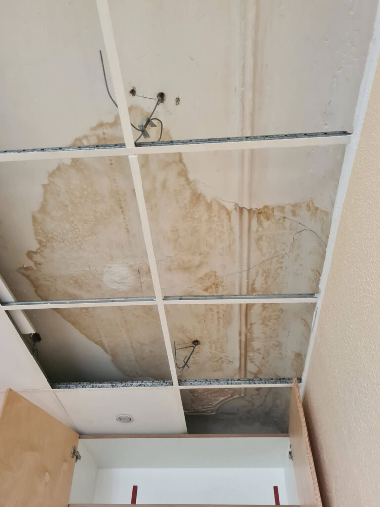 Eine decke mit fehlenden fliesen zeigt wasserbeschädigte, fleckige platten und freiliegende drähte, was auf ein mögliches leck oder ein strukturelles problem hinweist. einige schränke und wände sind an den rändern sichtbar.