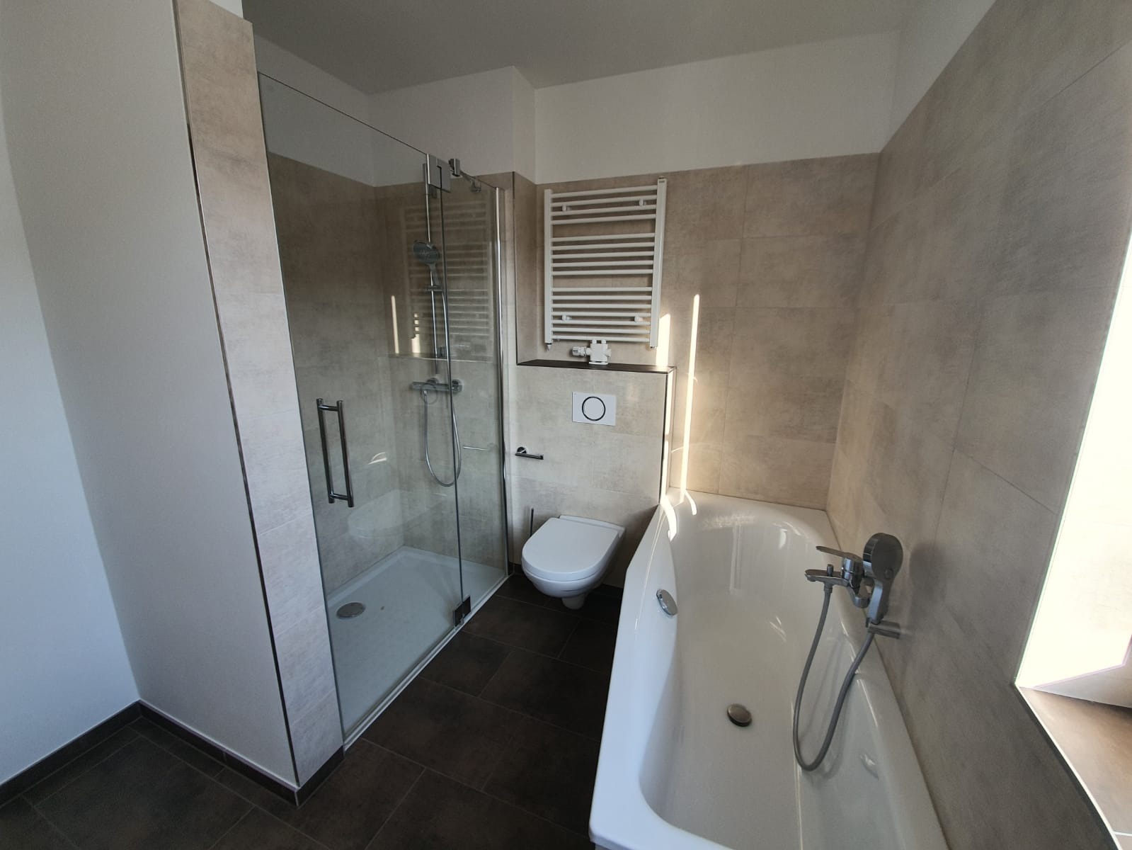 Modernes Badezimmer mit Glastür-Dusche, wandmontierter Toilette, weißer Badewanne mit Handbrause, Handtuchheizkörper und beigefarbenen Fliesen an Wänden und Boden. Natürliches Licht fällt durch ein Fenster neben der Badewanne ein.