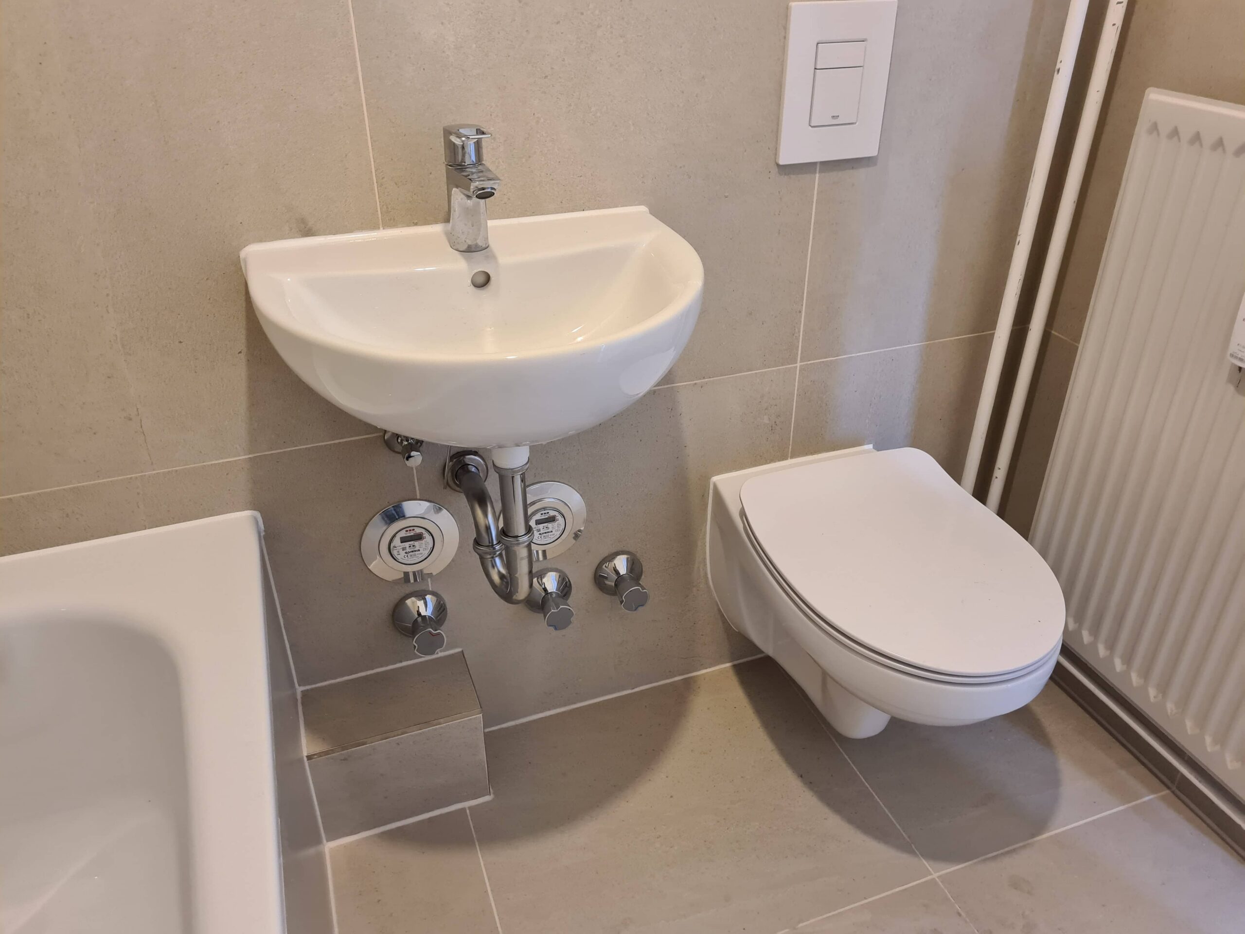 Ein kleines Badezimmer mit einem an der Wand montierten Waschbecken und Wasserhahn, darunter freiliegenden Rohrleitungen, einer wandhängenden Toilette, einem Teil einer Badewanne, beigen Fliesen, einem Heizkörper und einer Spültaste an der Wand.
