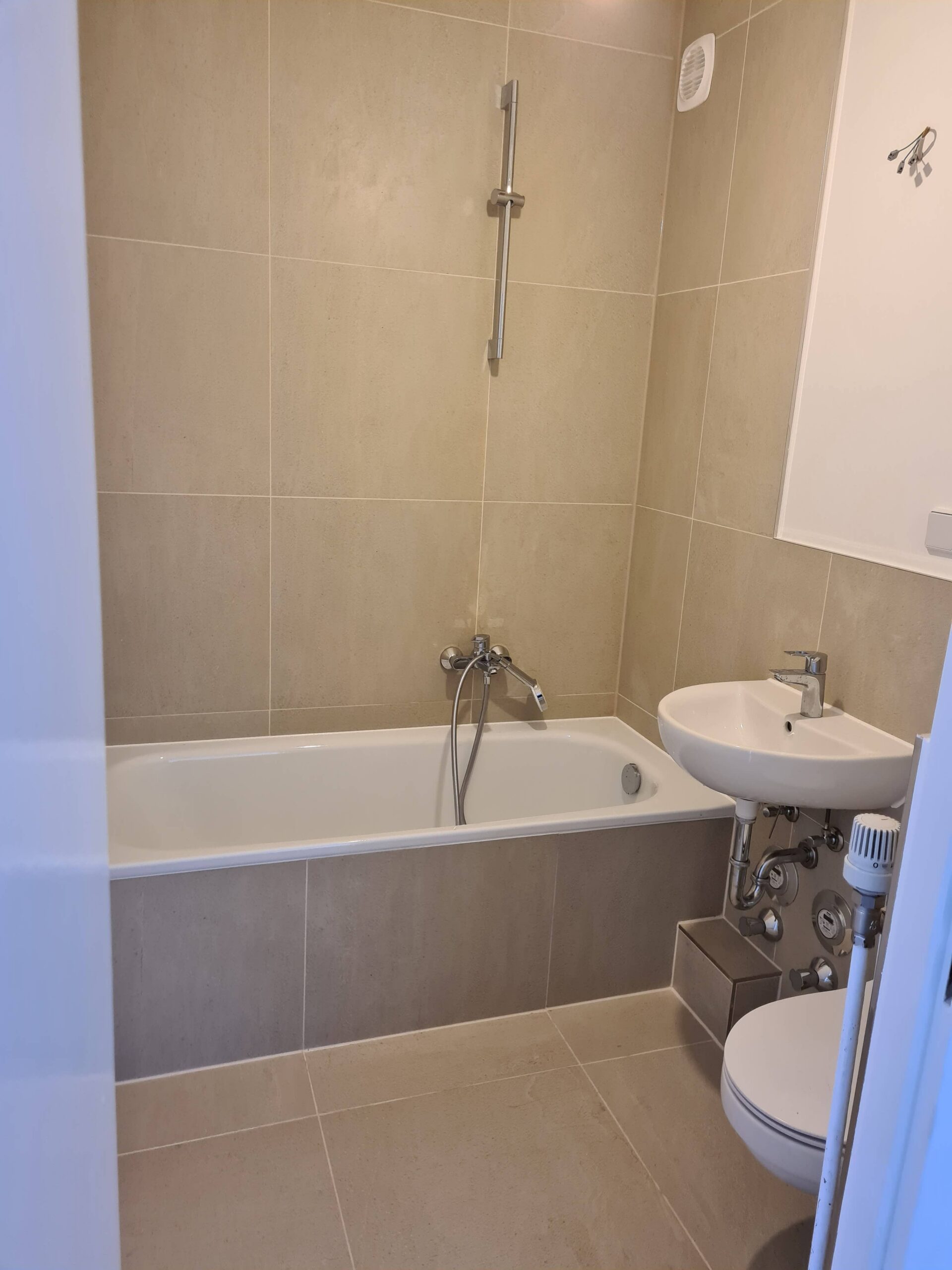 Ein modernes Badezimmer mit beigen Fliesen, einer weißen Badewanne mit wandmontierter Duschbrause, einem weißen Waschbecken mit freiliegenden Rohrleitungen und einem Teilblick auf eine Toilette. Die Wände und der Boden sind gefliest.