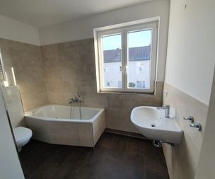 Ein modernes Badezimmer mit einer Eckbadewanne, einem an der Wand befestigten Waschbecken und einer Toilette. Es gibt ein großes Fenster, das natürliches Licht hereinlässt, beige geflieste Wände und einen dunkel gefliesten Boden.