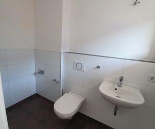 Ein minimalistisches Badezimmer mit weiß gefliesten Wänden, einer Wandtoilette, einem kleinen Wandwaschbecken mit Wasserhahn und einem dunklen Bodenbelag. Der Raum ist durch natürliches Licht hell erleuchtet.