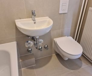 Ein kleines weißes Wandwaschbecken mit freiliegenden Rohrleitungen steht neben einer modernen Wandtoilette in einem gefliesten Badezimmer. Ein Teil einer Badewanne und ein Heizkörper sind ebenfalls sichtbar.