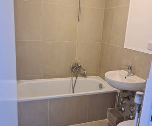 Ein kleines Badezimmer mit beige gefliesten Wänden, einer weißen Badewanne, einem Wandwaschbecken mit freiliegenden Rohren und einem silbernen Duschkopf über der Wanne.