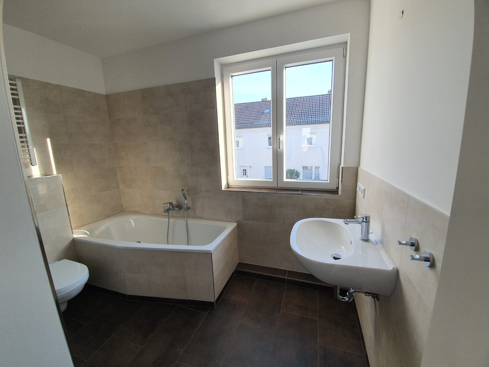 Ein modernes Badezimmer mit beige gefliesten Wänden, dunklem Fußboden, einer Badewanne mit Dusche, einem an der Wand befestigten Waschbecken, einer Toilette und einem großen Fenster, das Tageslicht hereinlässt.