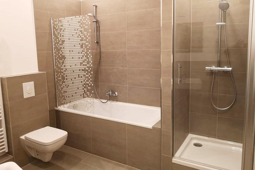 Modernes Badezimmer mit beigen Fliesen, einer Wandtoilette, einer Badewanne mit Dusche und dekorativer Glasabtrennung sowie einer separaten Dusche mit Glasabtrennung und Regenduschkopf.
