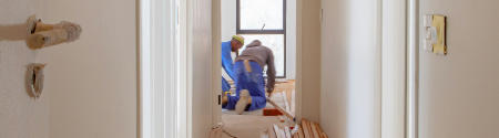 Zwei Arbeiter in blauen Overalls sind dabei, in einem Raum am Ende eines Flurs einen Fußboden zu verlegen oder zu reparieren, wobei Werkzeuge und Holzbretter auf dem Boden verstreut liegen. Die Wände des Flurs sind unbearbeitet.