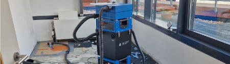Ein blauer Industriestaubsauger oder eine Reinigungsmaschine mit Schläuchen steht in einem Raum mit teilweise entferntem Bodenbelag, freiliegenden Rohren und natürlichem Licht, das durch große Fenster einfällt.
